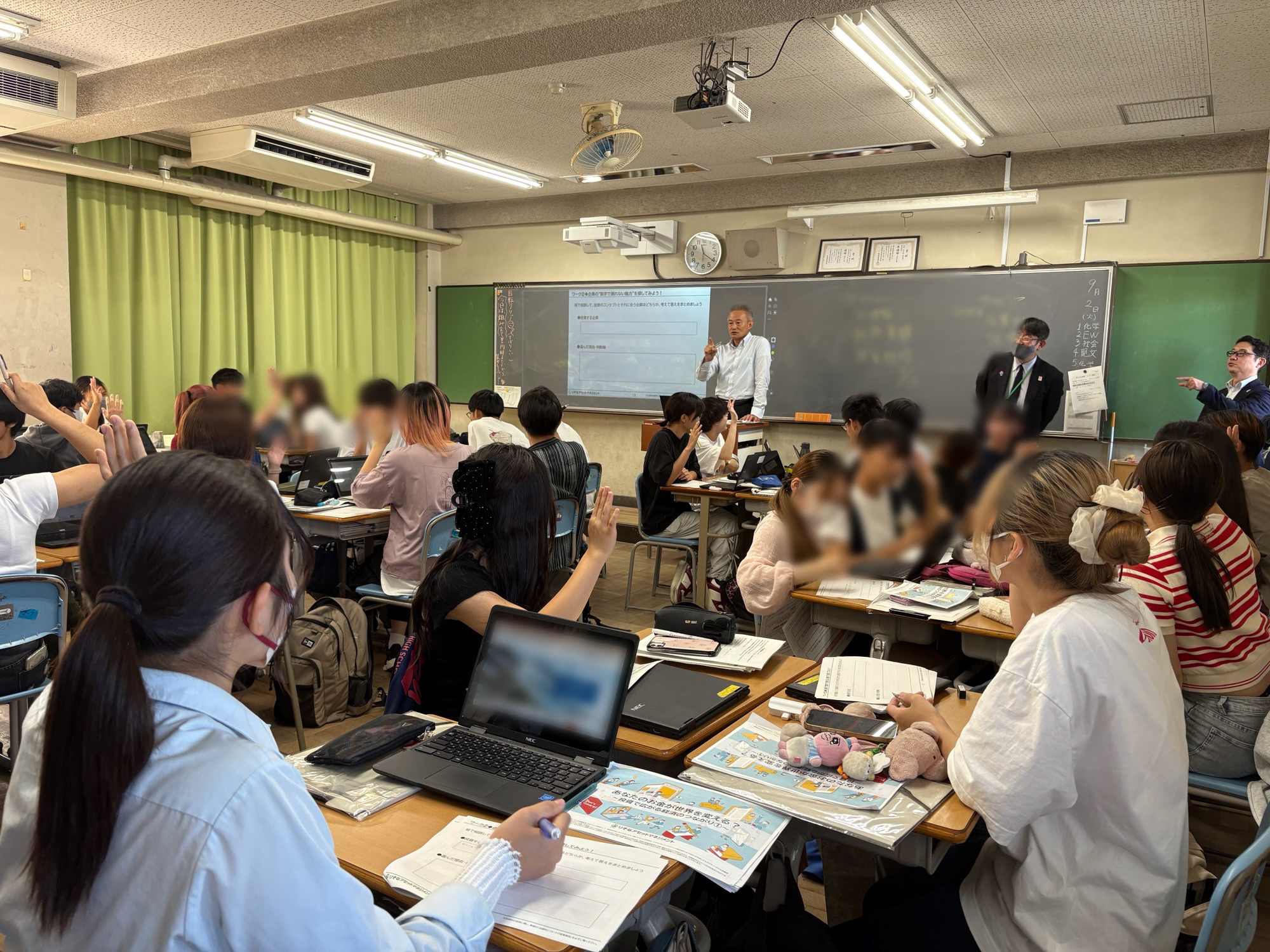 講師が生徒たちに、どちらの企業に投資するかを尋ね、手を挙げてもらっている画像